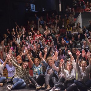 Foto del encuentro. Se ven muchas personas levantando manos en un auditorio