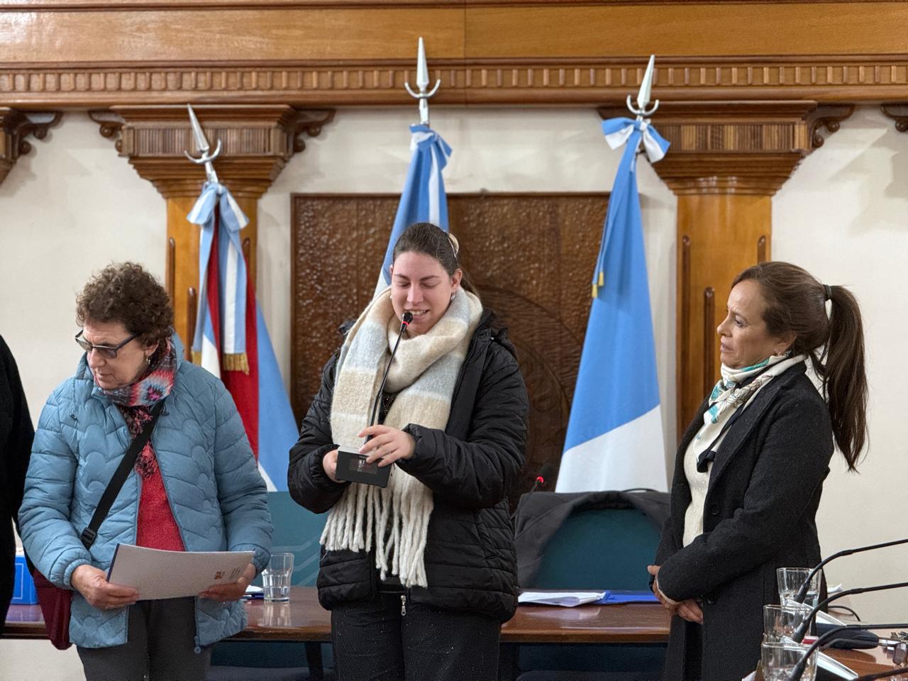 SE observa de fondo la sala de sesiones del Concejo Deliberante de Colón, con la bandera nacional y provincial. Delante, paradas, tres mujeres entre las cuales se encuentra Belén Noir luego de recibir el reconocimiento